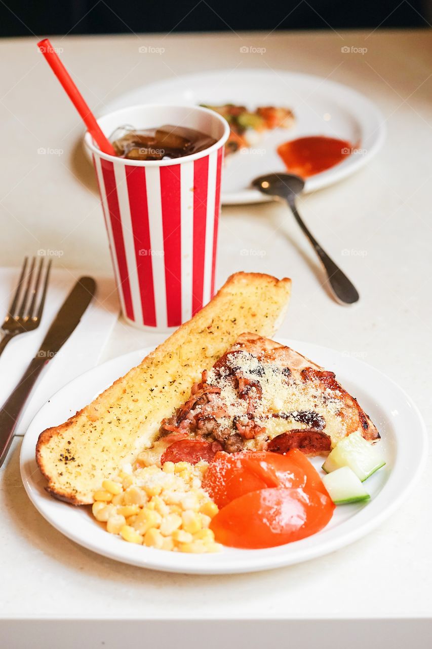 Fast food. Garlic bread and a slice of pizza with extra cheese, tomatoes, cucumber and sweet corns with carbonated drink. Selective focus on the pizza