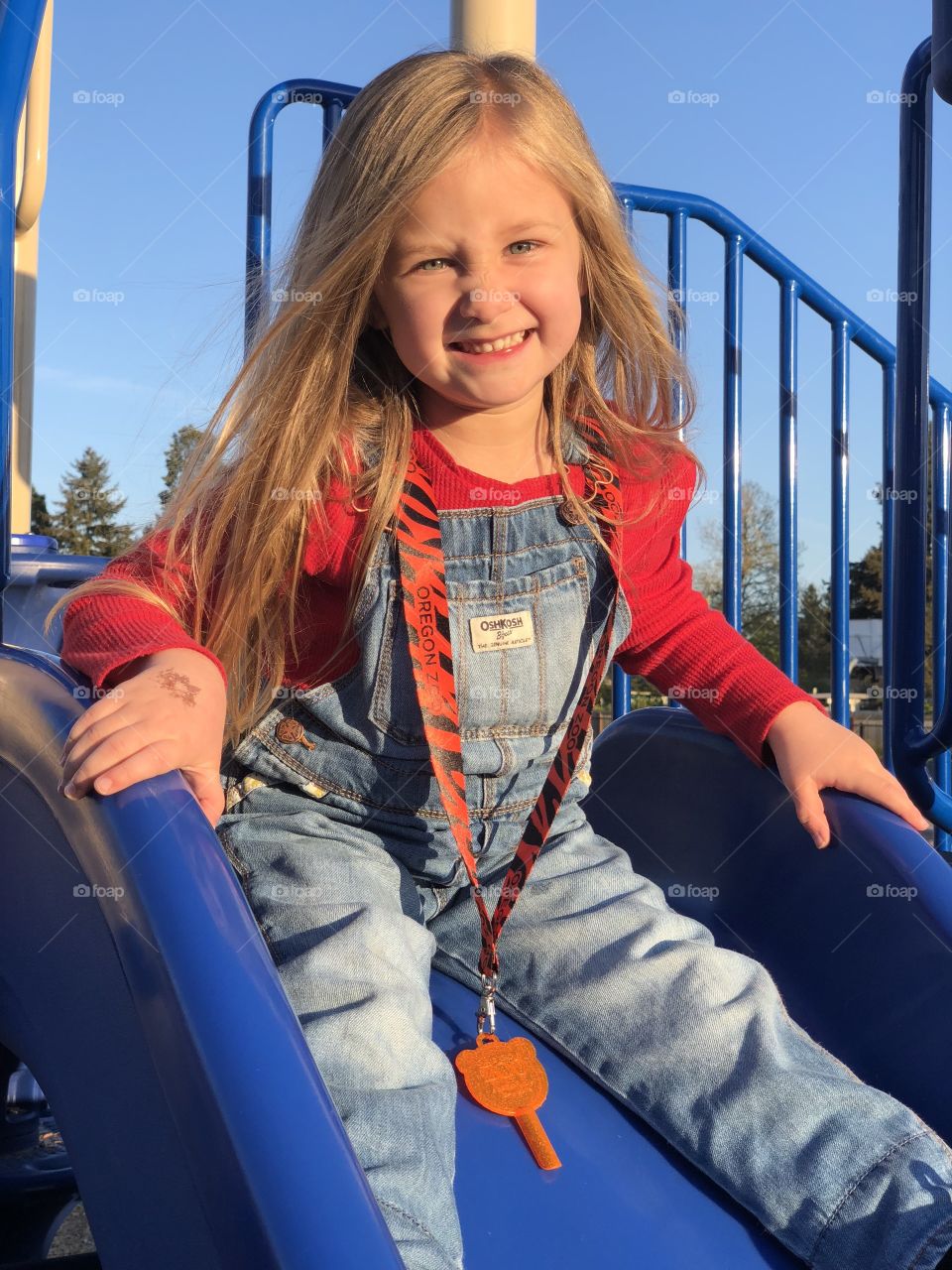 Little girl with blue eyes and long blond hair wearing overalls going down a blue slide at the playground