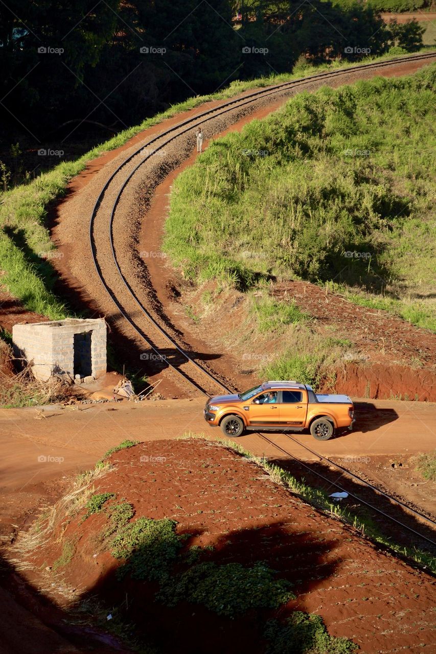 An orange pickup truck crossing a railway, in a semi-urban area, on a sunny afternoon. 