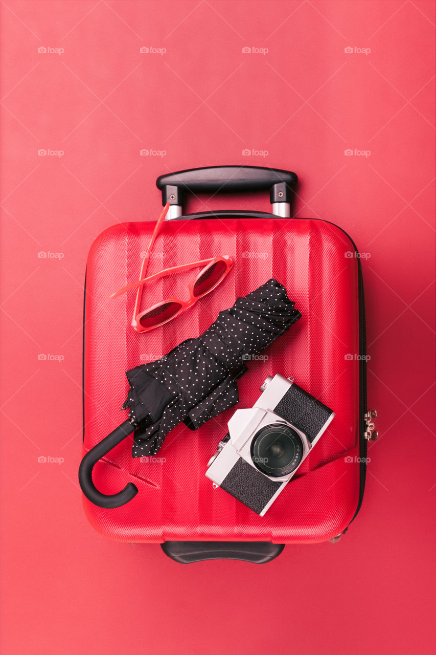 Red travel suitcase with red sunglasses, old camera, black umbrella on red background. Minimal style