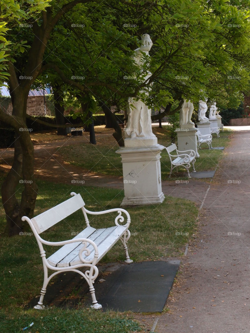 Outdoor benches in an urban park amongst amongst statues and next to a walking path on a pleasant summer day. 