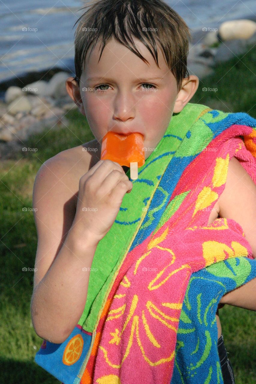 Young Boy with Colorful Beach Towel Eats Orange Popsicle on a Sunny Summer Day