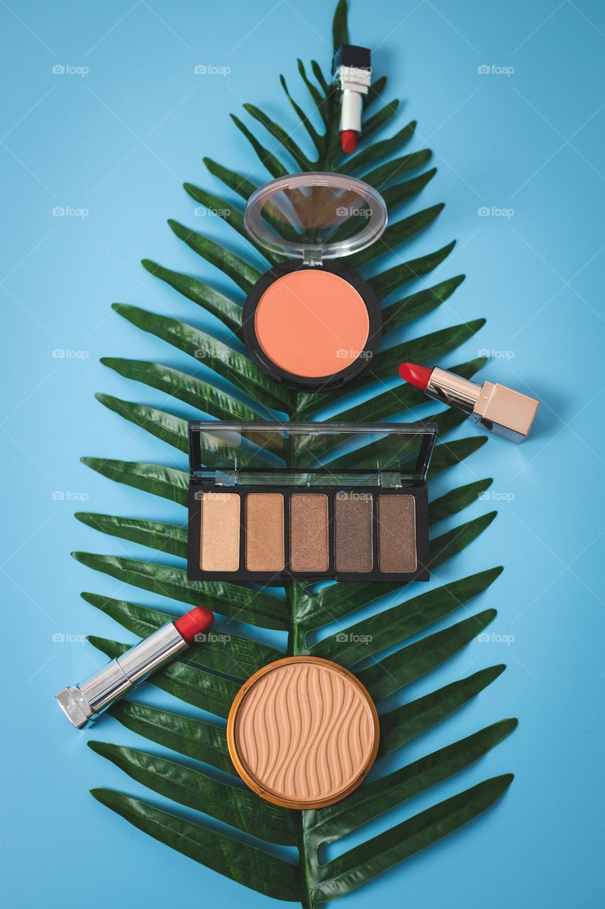 Eye shadow, powder, foundation and three lipsticks lie on a green palm leaf in the middle on a blue background, flat lay close-up.
