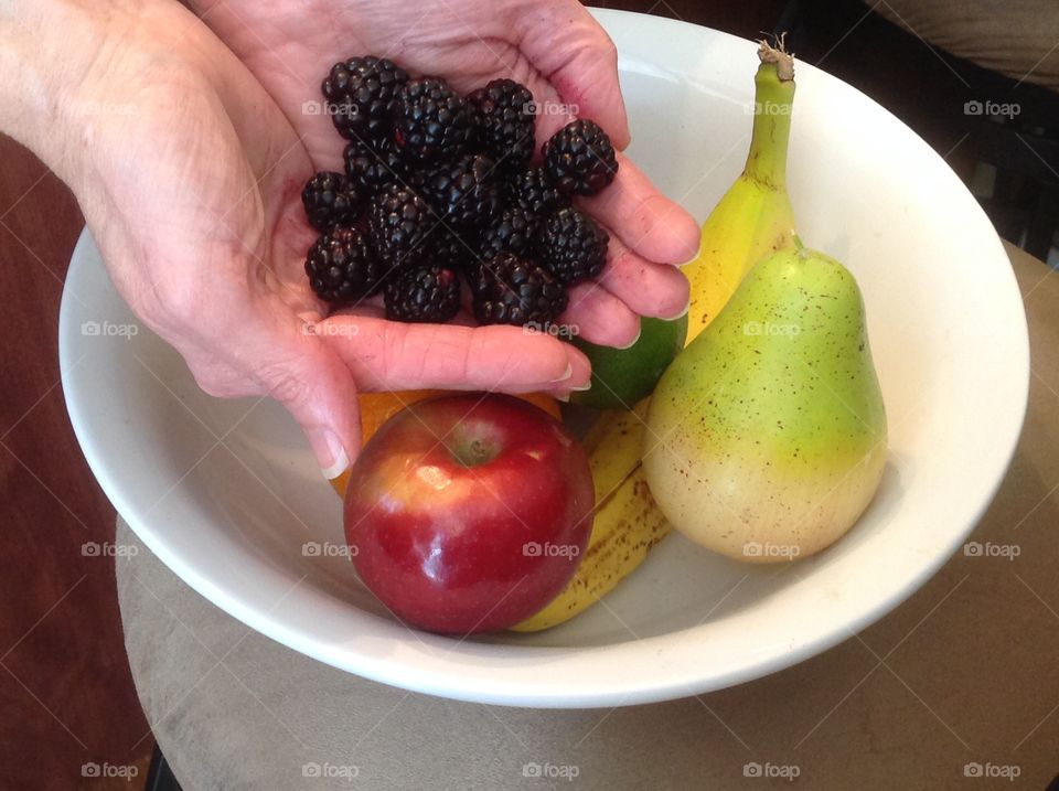 Holding in hand blackberries near a bowl of colorful fruit.