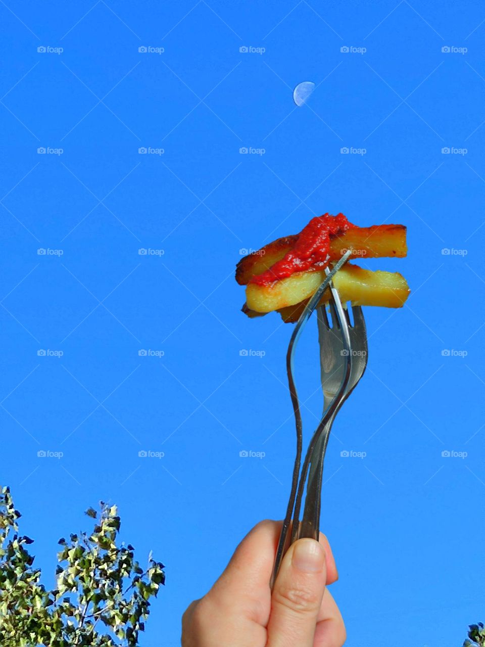 Food.  Dinner.  A hand against a blue sky holds three metal forks.  Pieces of fried potatoes with red ketchup lie on the tines of the forks.  The moon is visible in the sky