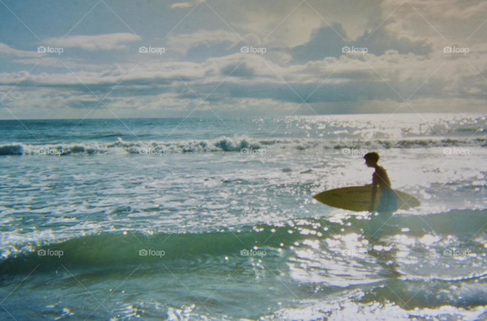 Boy carrying his surfboard into the water. Analog print photographed with my current camera 