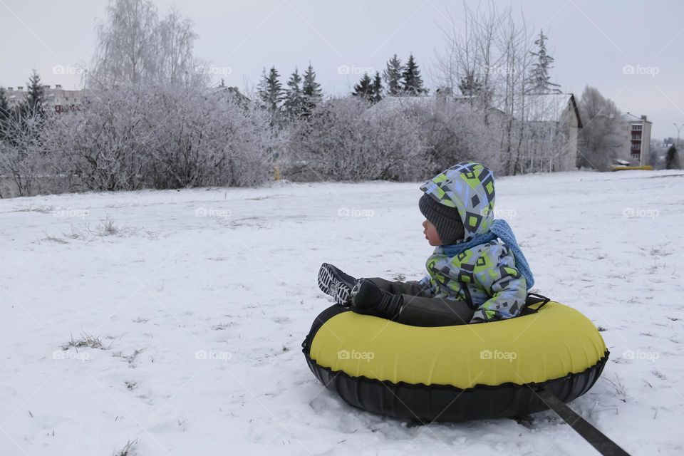 A small, carefree boy walks in the white snow in winter and rides a tubing in the park, near trees in the snow.