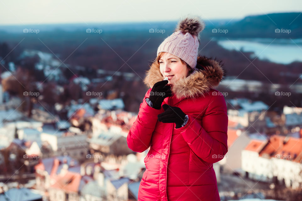 Woman applying lip balsam while walk on a wintery day. Wearing red coat and cap. Town in the background