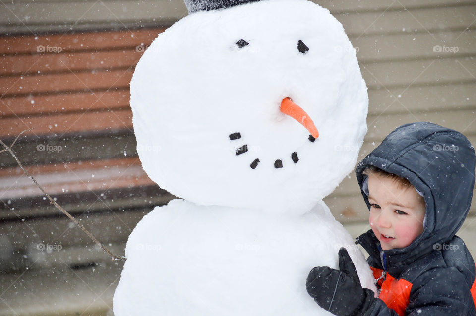 Toddler boy hugging a large snowman outdoors in the snow during winter