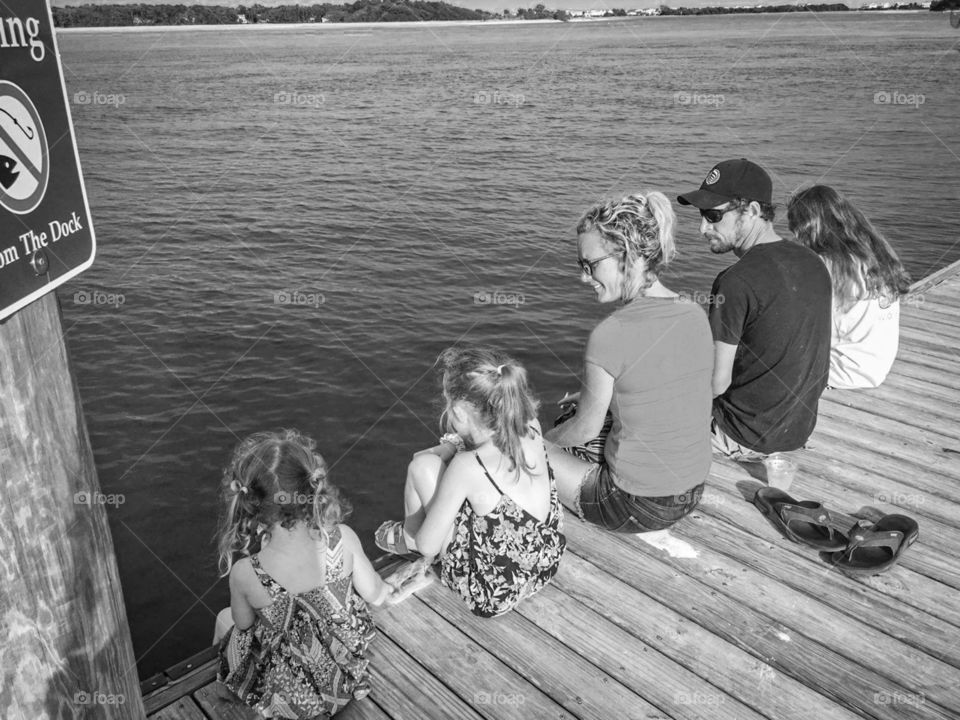 A family sits on a dock overlooking a body of sea water at the beach.