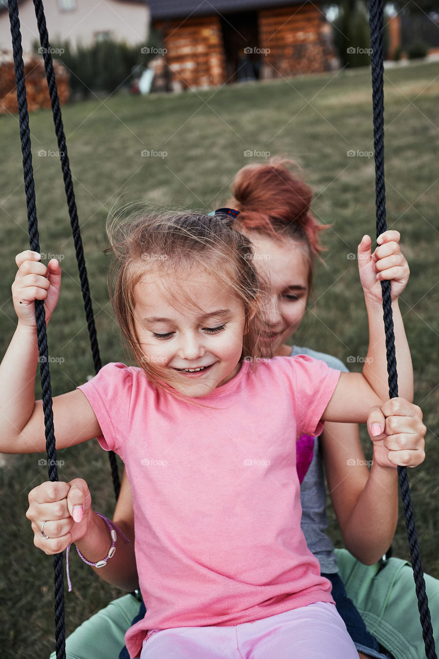 Teenage girl playing with her younger sister in a home playground in a backyard. Happy smiling sisters having fun on a swing together on summer day. Real people, authentic situations