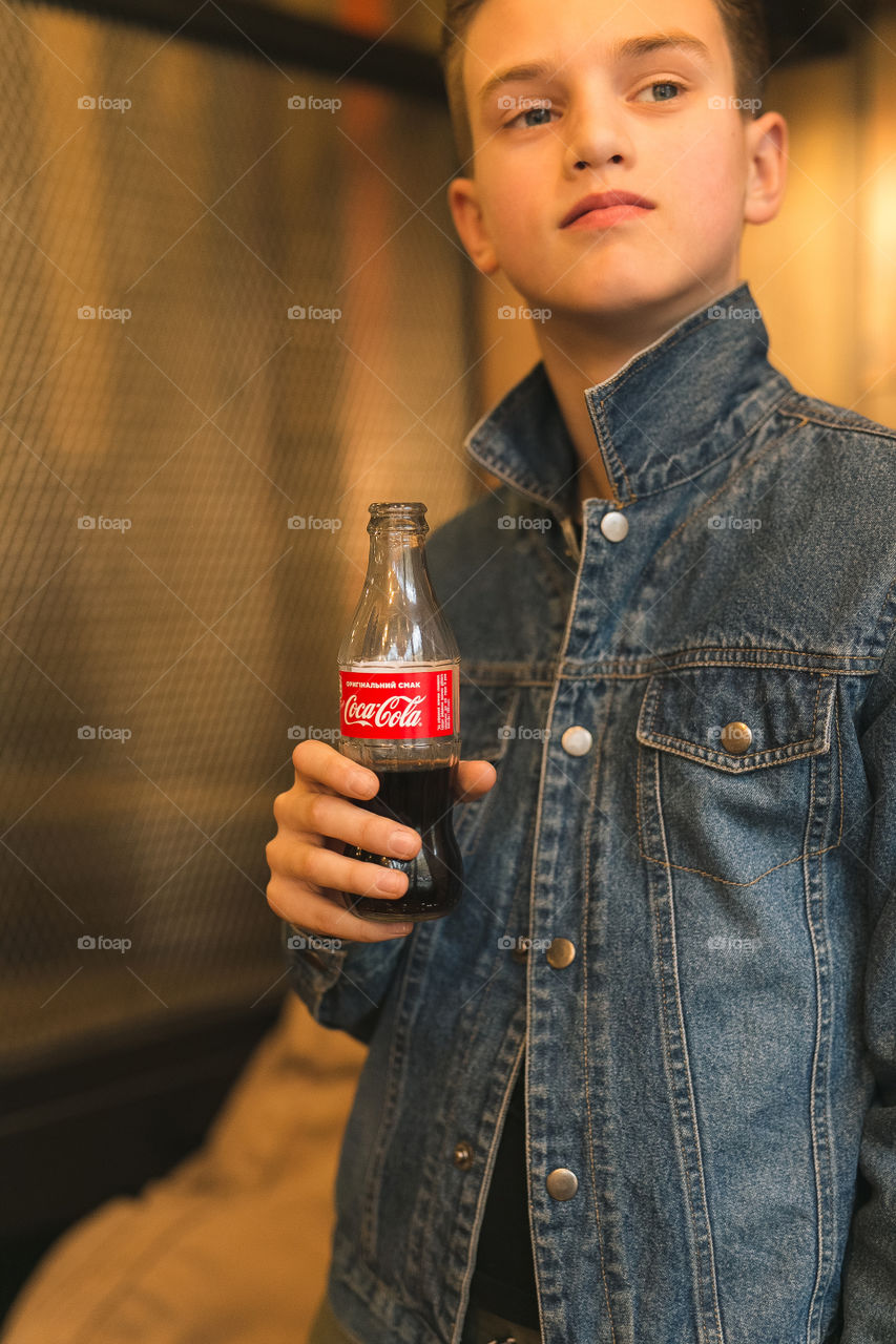 handsome guy, teenager, in a cafe, stands by the window, in a denim jacket, holds a bottle of Coca-Cola in his hands, drinks it, spends time