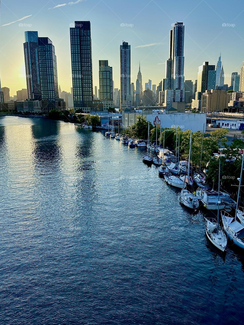 This is “Newtown Creek” with its many boats lined up side by side seen from the “Pulaski Bridge” that connects “Greenpoint”, Brooklyn to LIC, Queens. In the distance we see “Manhattan” including the “Empire State Building”. 2024. Hypnotic Productions