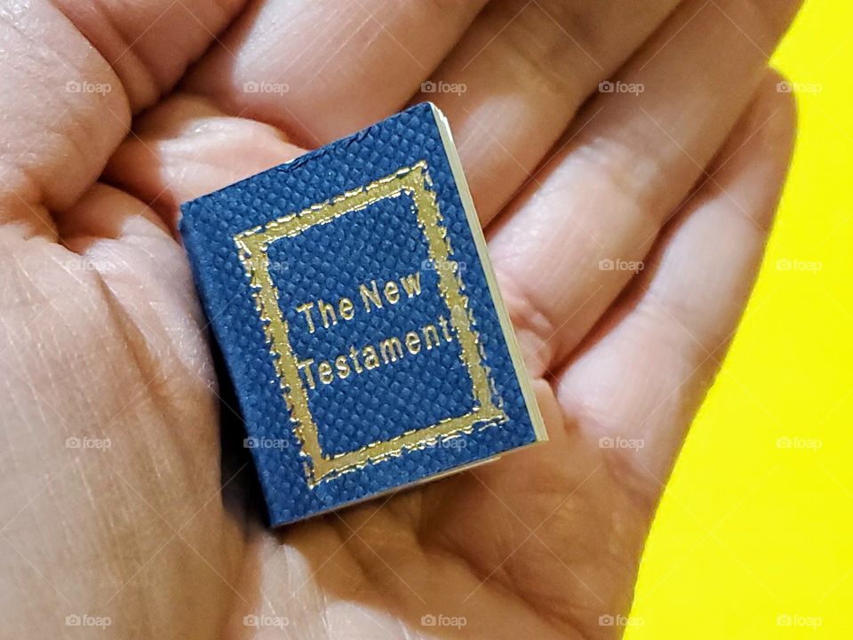 hand holding a miniature bible- the new testament. On a yellow background