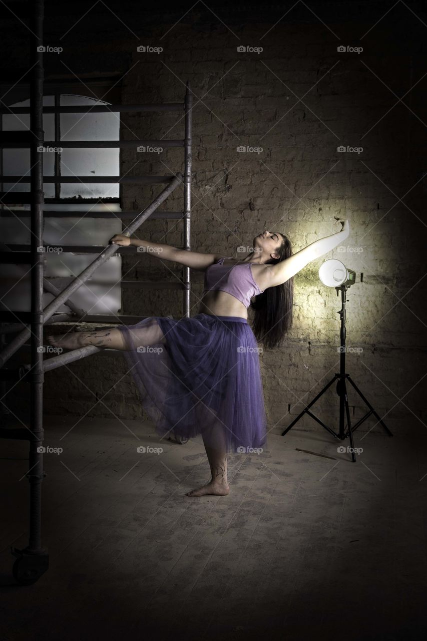 Girl dancing in a derelict building. Using scaffolding as a dance prop. Lit from behind with studio light. Wearing purple skirt and crop top. Leaning back in a dance position whilst holding on to metal scaffolding