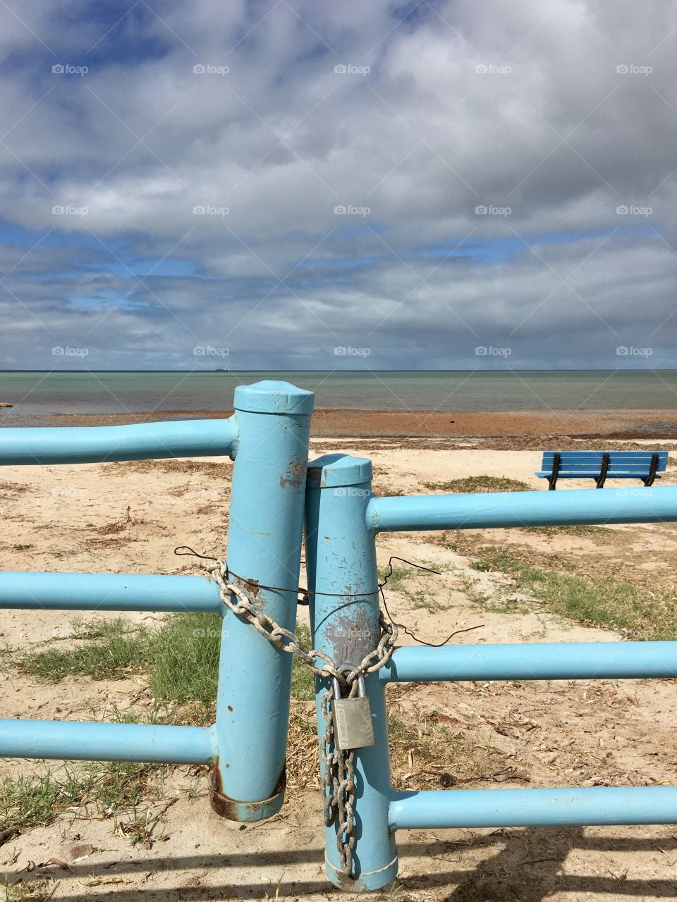 Aqua marine blue metal gate locked with padlock and chain as a barrier to vehicles entering the beach, blue bench and ocean horizon in the background