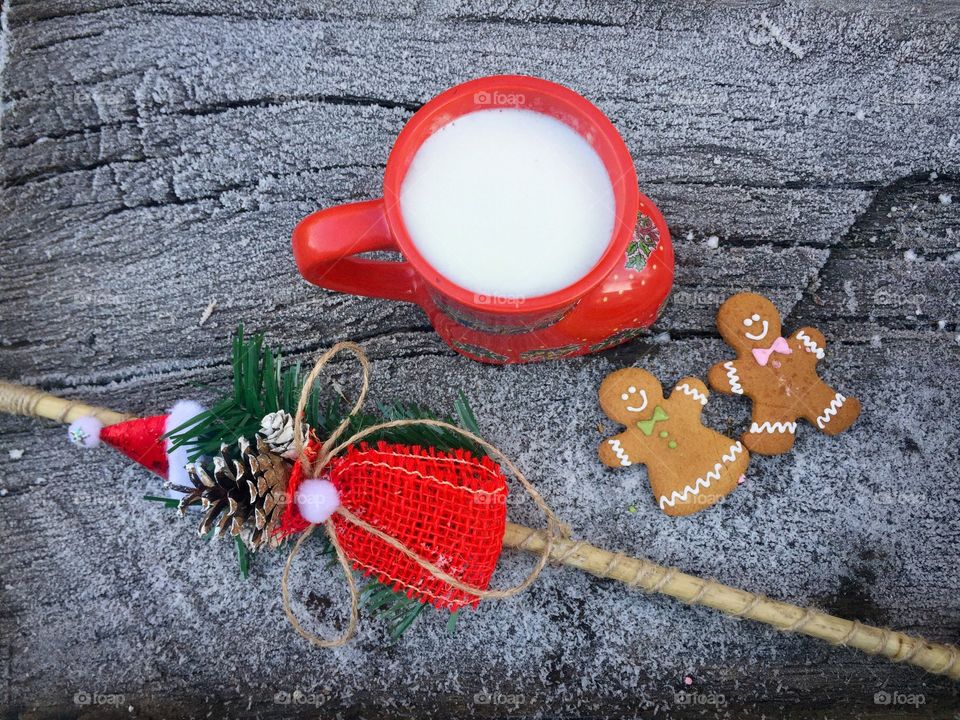 Red boot-shaped mug of white chocolate and milk  on snowy table with two gingerbread men beside and frozen pine cone tree branches 