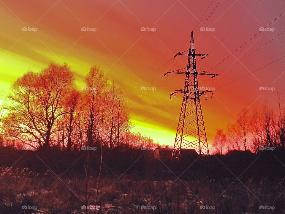 осенний вечер, закат, силуэт деревьев на фоне оранжевого неба, тёмные облака, вышка линии электропередачи.