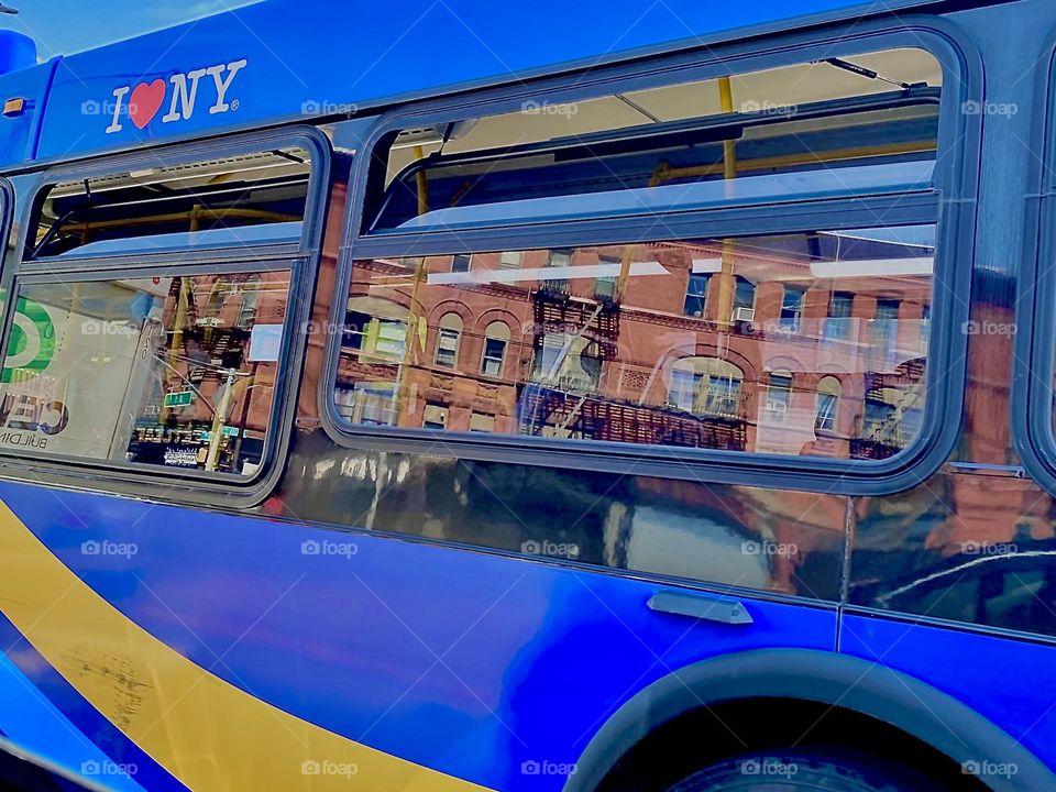 “I love NY” says the inscription on this indigo blue bus driving past us in “Fort Greene”, Bklyn. Several old style brownstones are reflecting in the large bus windows from across the street. 2022. Hypnotic Productions