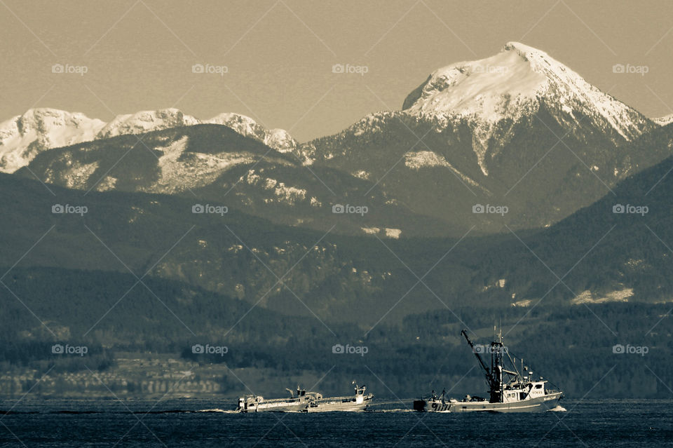 Split-tone monochrome. Landscape of a herring fishing boat pulling 2 haulers along the strait. Shades of blue-grey & grey provide additional contrast between sea, shore, hills, mountains & sky.