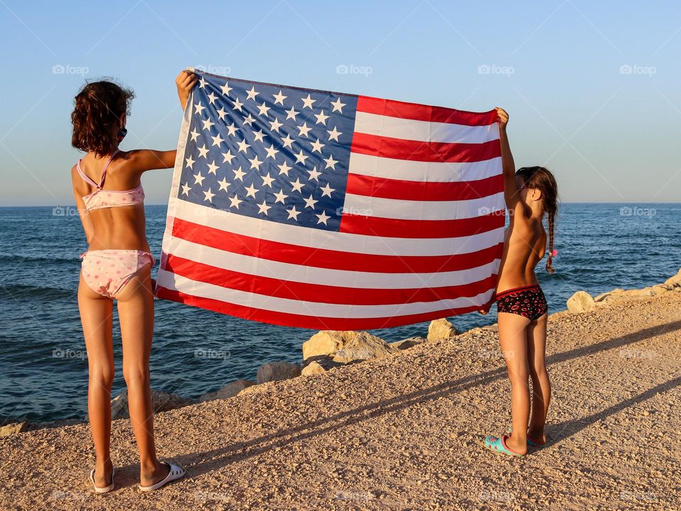 Two Caucasian girls in swimsuits holding an American flag on the seashore at sunset hour, close-up view from the side.