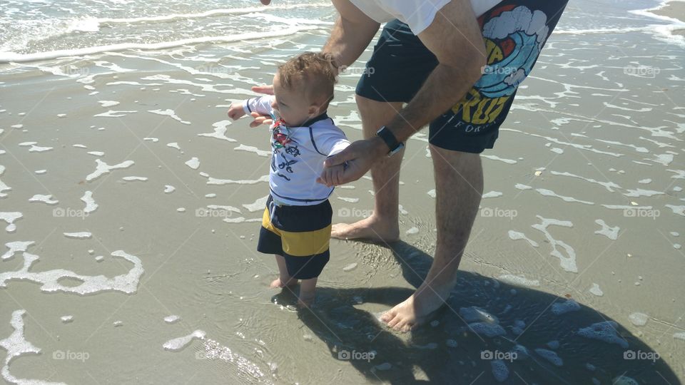 A baby boy stands in the ocean for the first time with his dad holding his hands