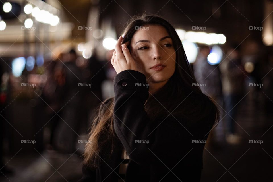 Portrait of a beautiful young caucasian girl with her hand straightens her hair behind her ears while walking in night Prague, close-up side view. Night walk concept.