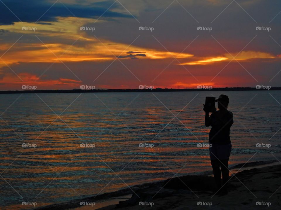 A lady standing on the shoreline of the bay and taking pictures of the blended sunset with her iPad