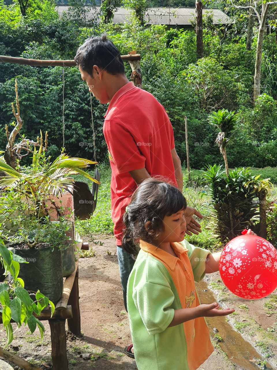 a father in a red shirt is looking at flowers in the yard with his daughter holding a balloon