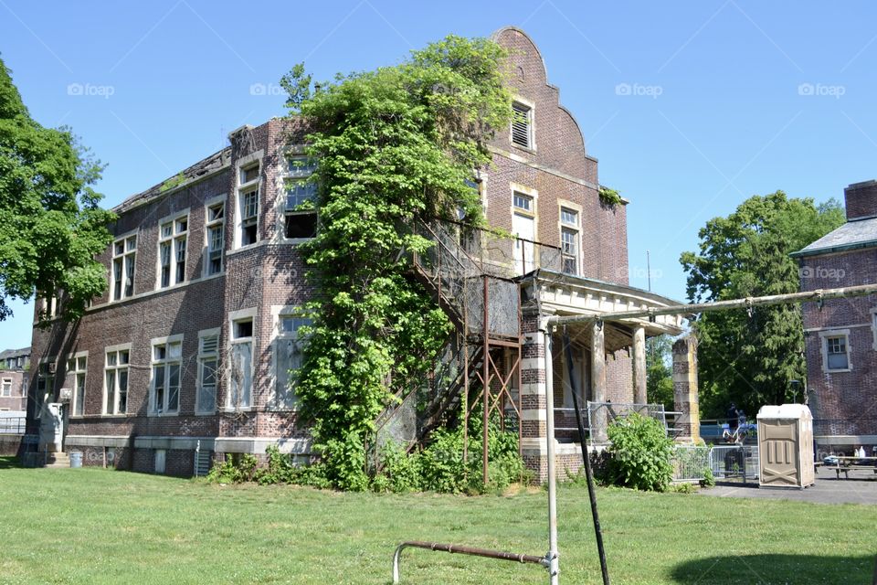 A partial old metal playground in front of and old abandoned brick building that’s overgrown with vegetation 