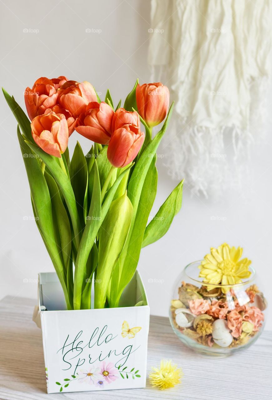 Orangey-red tulips in box labelled spring, with a bowl of pot pourri and white scarf hanging in the background 