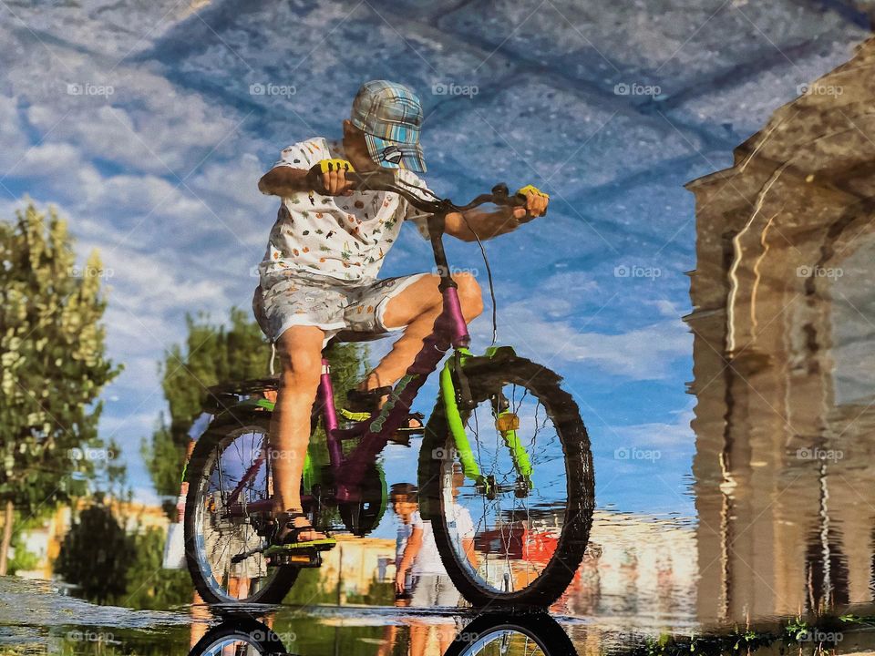 A kid on a bike reflection in a puddle during summertime playtime