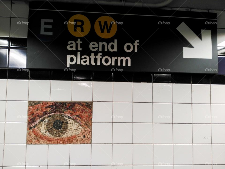 Green eye made of Tiles on a white tiled wall below the sign indicating the platforms in a New York subway station.