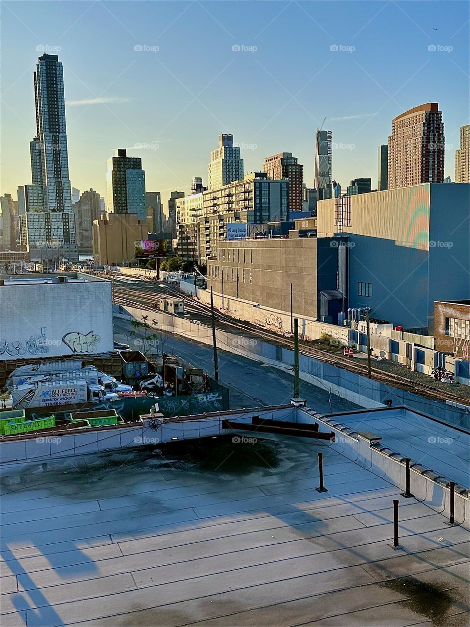 This is the top of the red metal staircase of the “Pulaski Bridge” at “Newtown Creek” in LIC, Queens overlooking the “Long Island Railroad” tracks, “iFly”, an indoor flight simulator and in the distance “Manhattan”. 2024. Hypnotic Productions