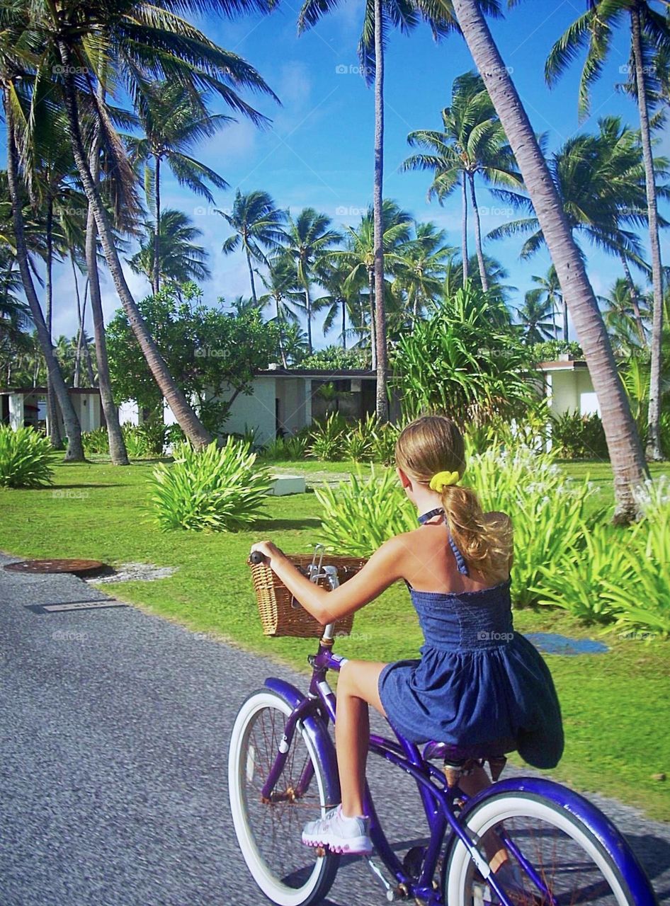 A young girl rides her bright purple bike on an island road, vibrant green palm trees move in the breeze ahead