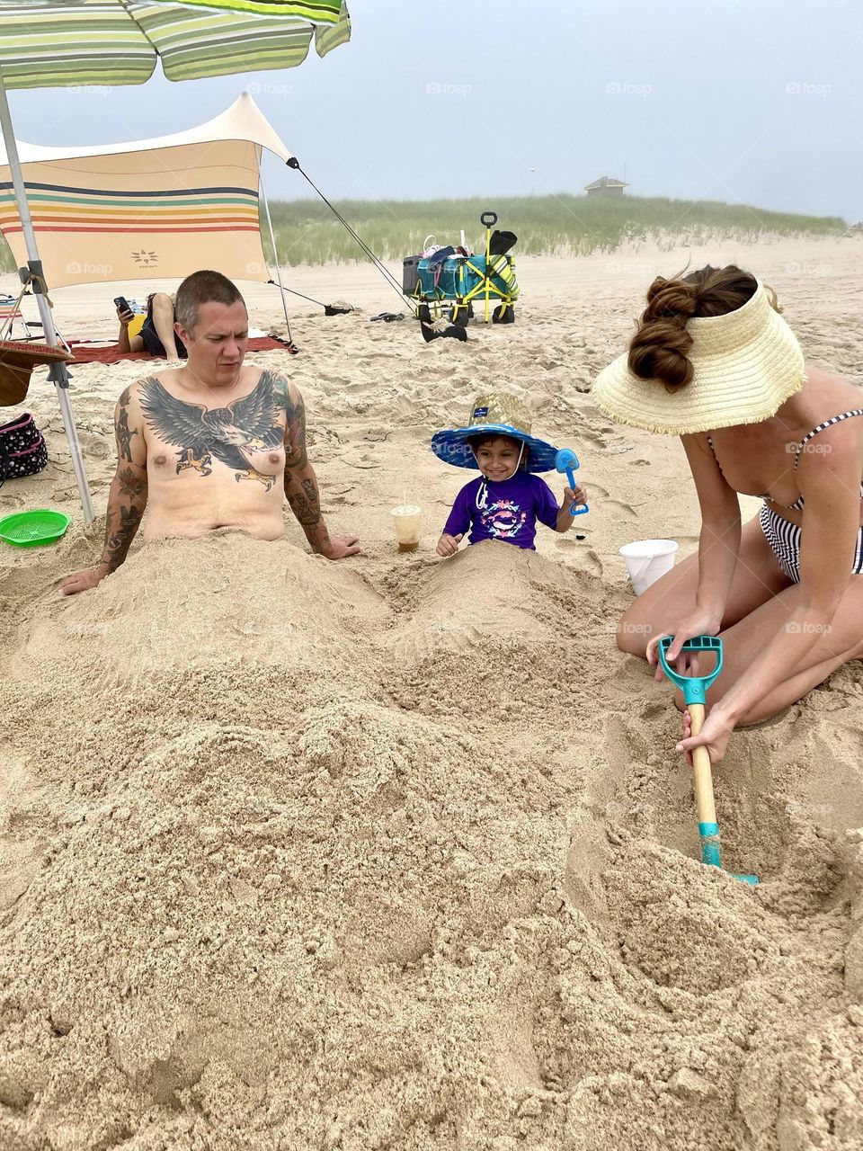 Family at the beach getting buried in the sand, funny sand stunts, making mermaids in the sand, little girl has fun in the sand, sand stunts in the summertime