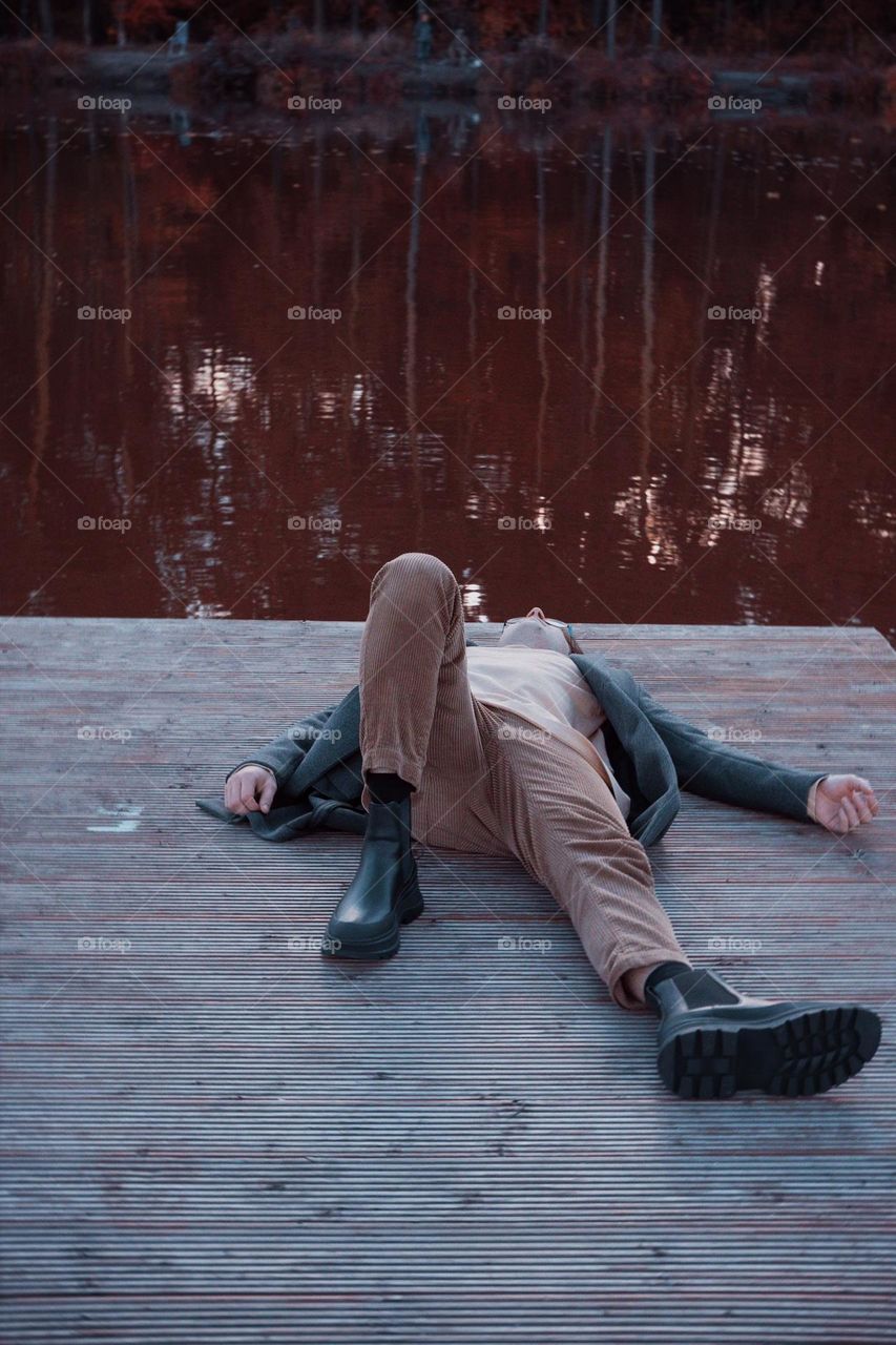 A man in a raincoat lies on a bridge against the background of a lake
