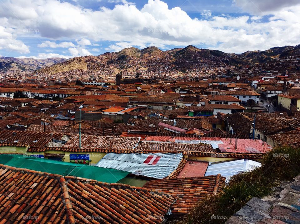 A view of Cuzco from a hill in the city shows colorful rooftops and mountains and clouds in the distance. 