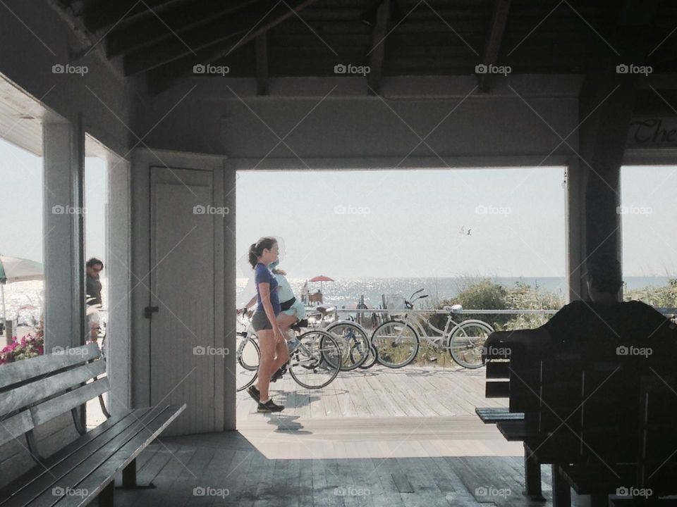 People walk and ride bicycles on the Ocean Grove boardwalk, photographed from the open-sided Beach Pavilion, where church services are frequently held. The Pavilion offers shade and a beautiful view of the ocean, beach and passerby.