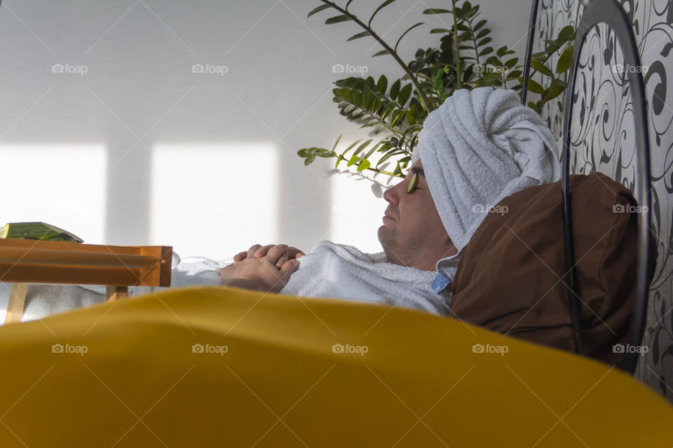 A man and home spa care at home takes care of himself and smears his face with cream, does a manicure at home on a blue background and with oranges and rests.