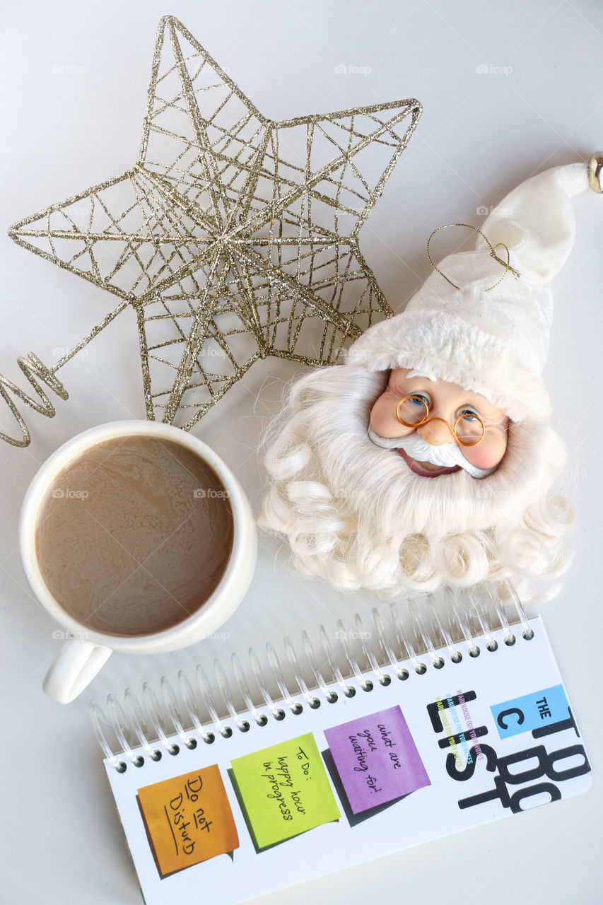 Overhead shot of a new year based scene. Notebook with a cover to do list written on, white Santa Claus ornament, coffee in a white cup, golden coloured star shaped christmas ornament.
