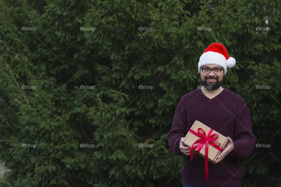 Smiling positive handsome hipster man in cozy red sweater santa claus hat holding a kraft gift tied with a red bow copy space. Concept: winter holidays weekend with family winter forest Black Friday