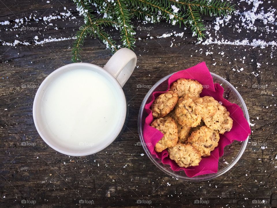 Mug of milk with cookies on wooden table powdered with snow and pine cone tree branches