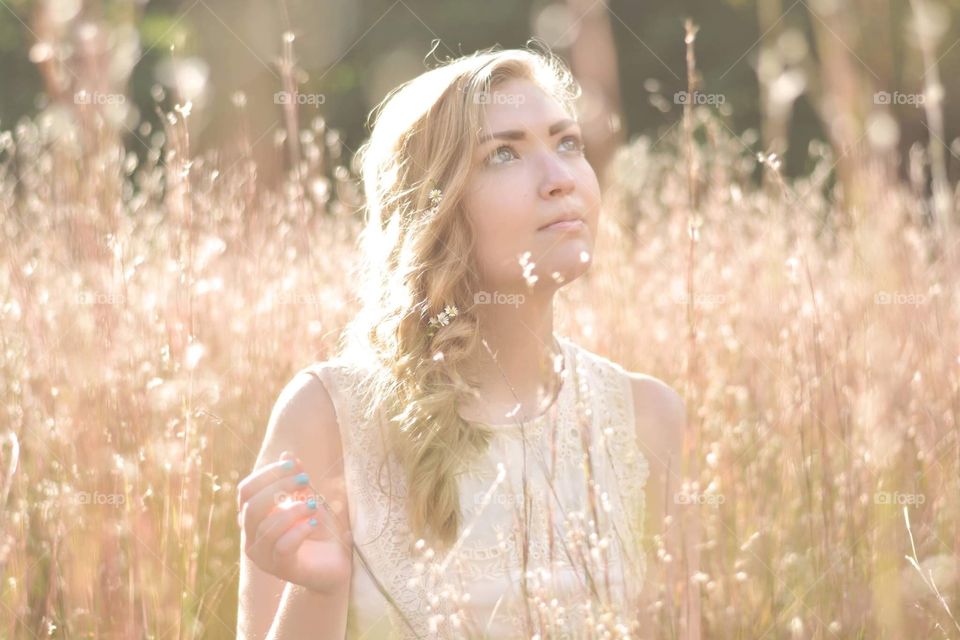 A young woman looks up in a golden field in summer in a white dress and flowers in her hair 