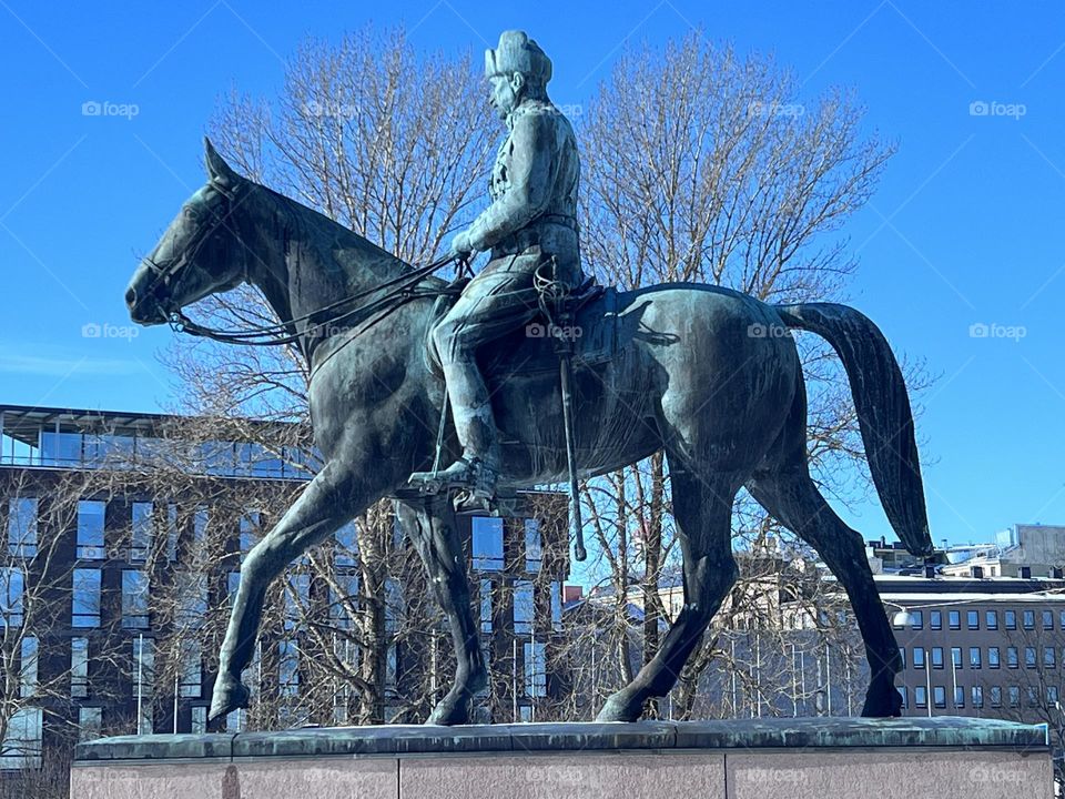 Close view of the bronze statue of the Finnish marshal Carl Gustaf Emil Mannerheim on the horse back against trees branches and deep blue sky