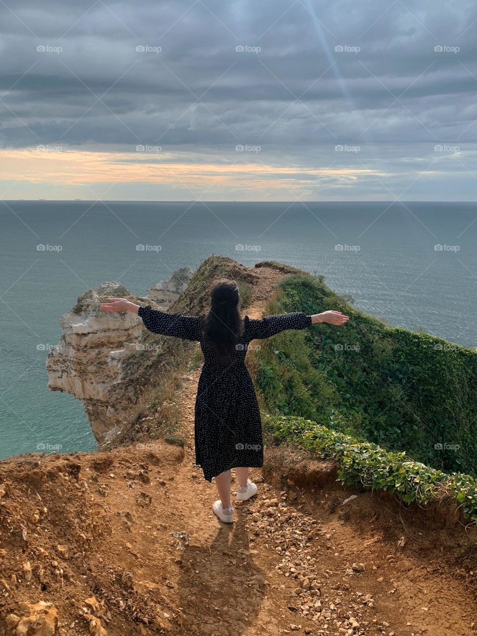 A young Caucasian girl in a black dress with white polka dots stands with her back on a high rock with her arms outstretched to the sky and inhales the aroma of the northern sea in Normandy in France, close-up side view.
Travel time concept, traveler