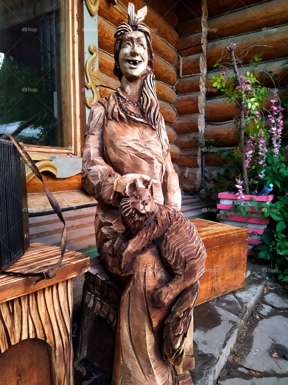 A figure that is carved out of wood.  Smiling old woman sits on a bench.  A cat is sitting on the lap of an old woman