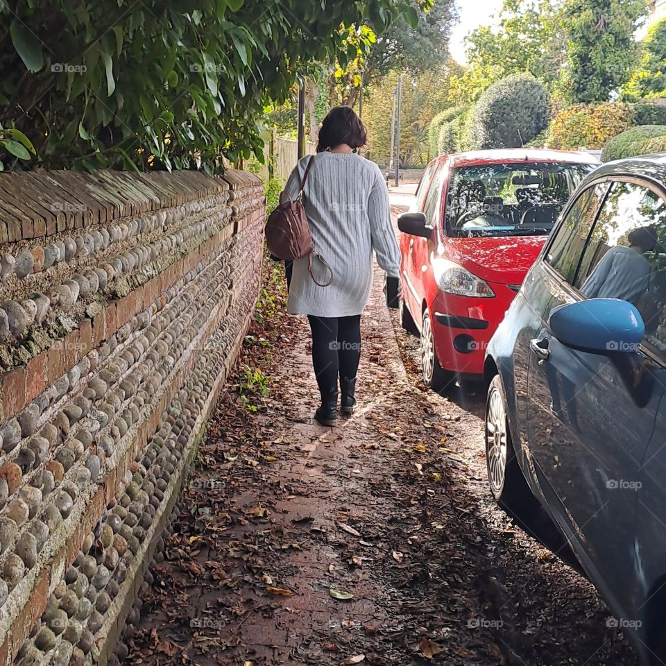 lady in jumper and boots walking down an autumn leaf strewn pavement with trees overhanging and parked cars. u.k