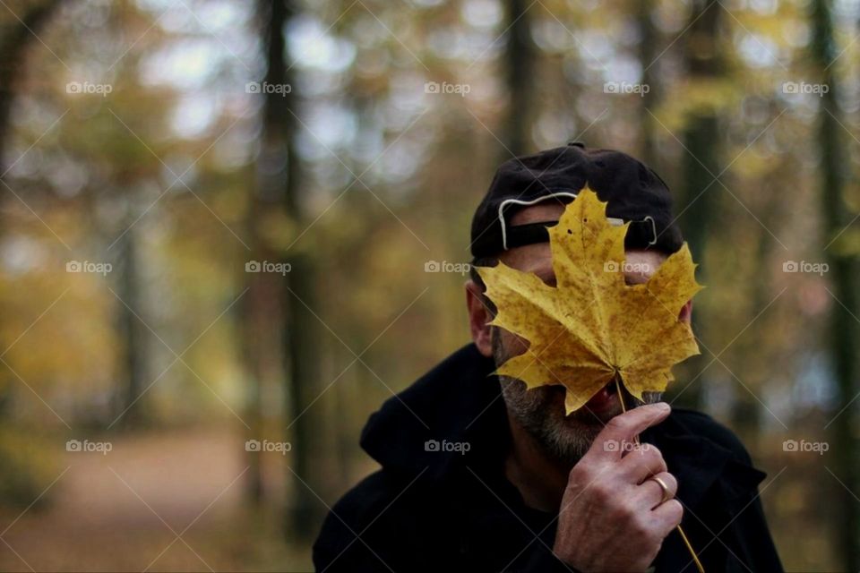 A bearded man with a black cap holds an autumn leaf to his face in the forest