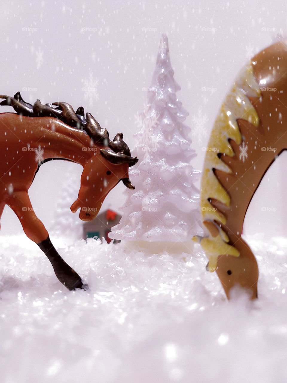 A beautiful magical story telling miniature white snow covered winter scene. A brown horse and a tan horse up front, a snow covered pine tree in the distance and a log cabin in the background with snowflakes falling everywhere.